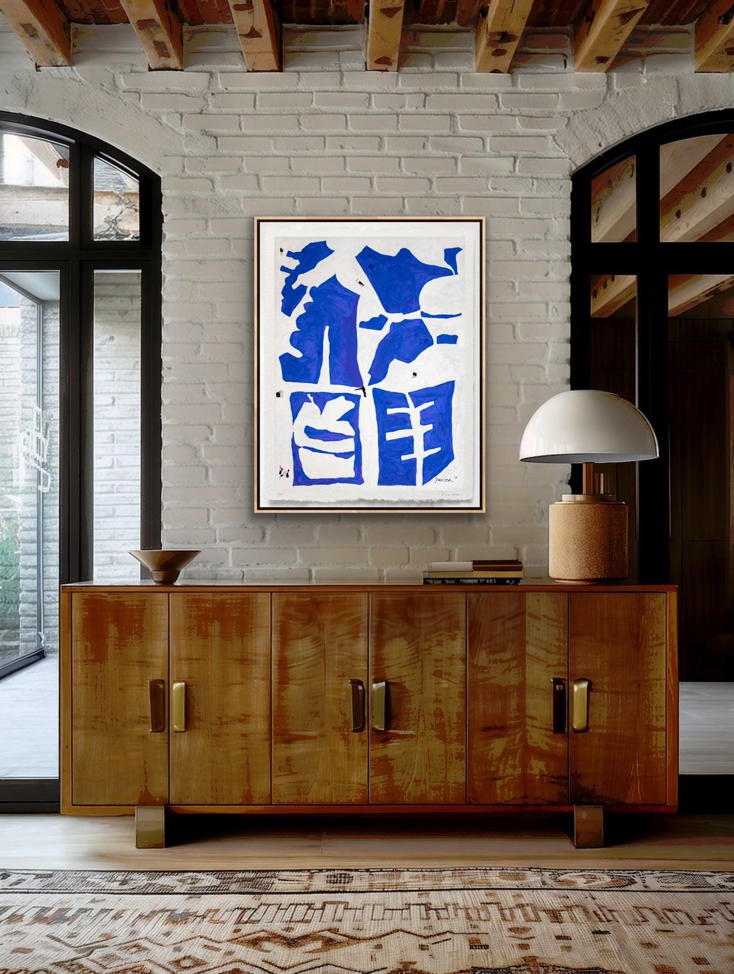 Wooden sideboard with a lamp and abstract blue artwork on a white brick wall.