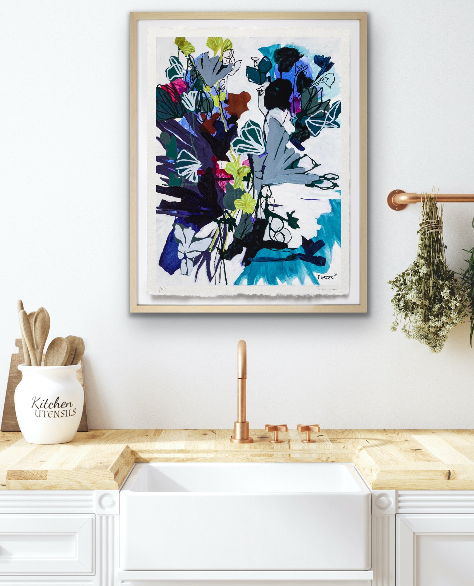 Framed abstract art above a kitchen sink with wooden cabinets and a white wall.